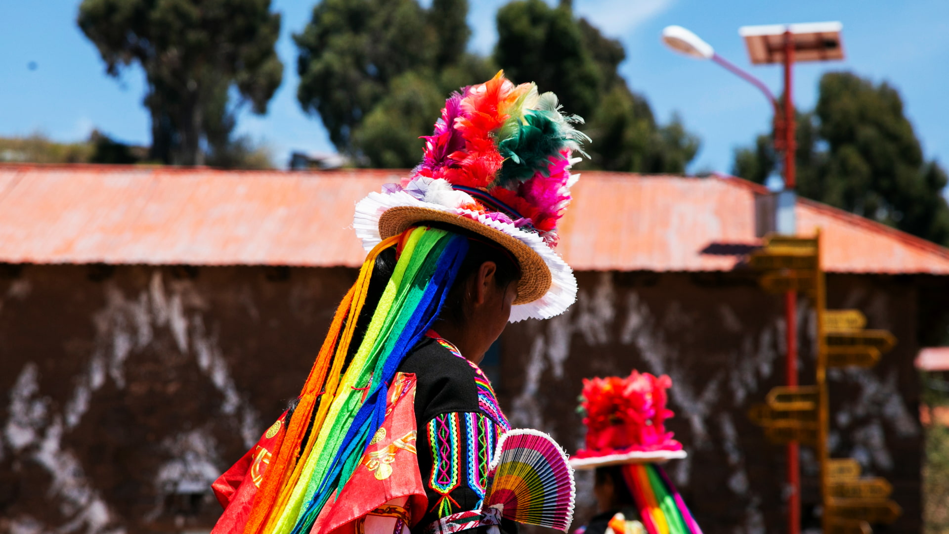 Danzante de Sikuris de espaldas, con un sombrero de plumas y cintas arcoíris, llevando un abanico. El vibrante traje de las danzas de la fiesta de Puno.