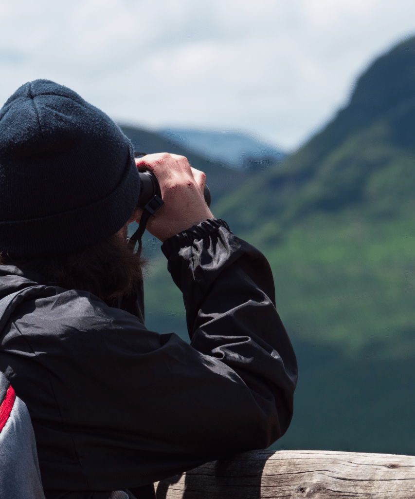 Viajero con casaca impermeable negra y gorro de lana usando binoculares frente a las montañas verdes, representando el equipaje para lluvia necesario en rutas de Cusco y Puno.